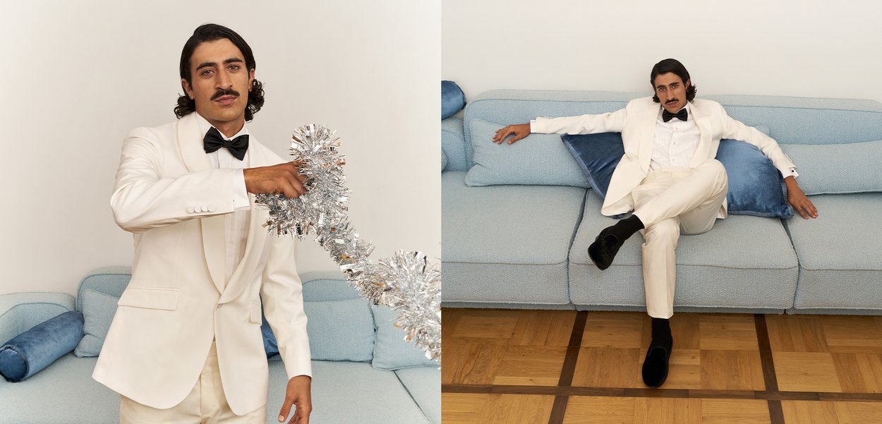 Fato branco com gravata borboleta preta, segurando papel prateado, sentado em um sofá texturizado azul claro com almofadas azul escuro; chão de madeira.