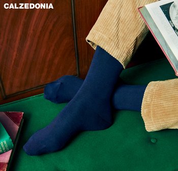 Person wearing navy blue socks and beige corduroy pants sitting on green velvet with books nearby and one book open.