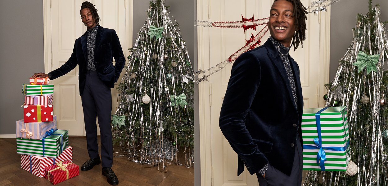 Mann in einem Samtblazer und gemustertem Rollkragenpullover steht neben einem geschmückten Weihnachtsbaum und hält verpackte Geschenke.