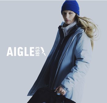 Long manteau bleu clair avec fermeture éclair à l'avant, superposé à une veste noire. Le mannequin porte un bonnet en tricot bleu et tient un sac noir.