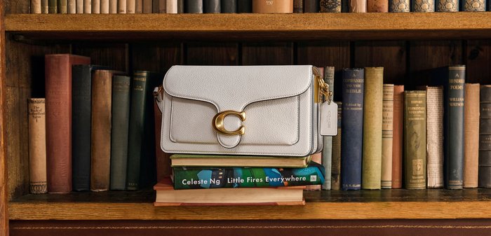 Weiße Leder-Schultertasche mit goldener Schnalle, die auf gestapelten Büchern in einem Bücherregal aus Holz liegt, das mit Vintage-Büchern gefüllt ist.