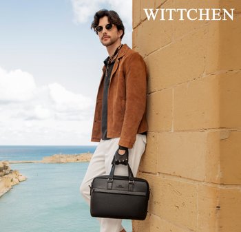 Mann mit Sonnenbrille und brauner Jacke lehnt an einer Steinmauer, hält eine schwarze Ledertasche und blickt unter bewölktem Himmel auf den Ozean.