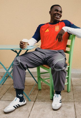 Άνδρας με κόκκινη αθλητική φανέλα και γκρίζα παντελόνια, καθισμένος σε πράσινη καρέκλα, κρατώντας ένα αναλώσιμο φλυτζάνι καφέ, χαμογελά και κοιτάει πλάγια.