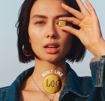 Moneda redonda de latón con "Lee" en relieve, sostenida contra la cara. El modelo lleva una chaqueta de mezclilla y pendientes de aro dorados, con un fondo neutro.