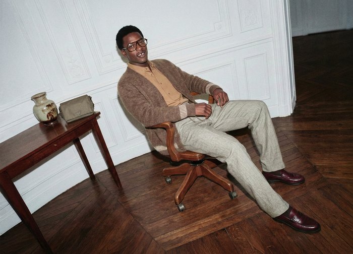 Homme portant des lunettes, chemise beige, cardigan marron et pantalon clair assis sur une chaise pivotante en bois sur un parquet foncé.