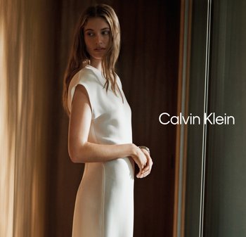 Mujer con vestido blanco sin mangas cerca de una ventana, mirando de lado con expresión serena, iluminación cálida interior, logo de Calvin Klein a la derecha.