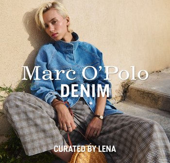 Blondynka w oversize'owej jeansowej kurtce i w kratkę spodniach siedzi na kamiennych schodach obok beżowej ściany, trzymając plecioną torebkę.