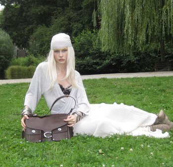 Brown leather handbag with silver hardware, featuring a flap closure, two front pockets, and a shoulder strap. Model in light gray outfit seated on grass.
