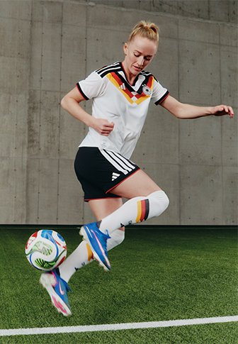 Weibliche Fußballspielerin im Deutschland-Trikot, die einen Ball in der Luft auf Kunstrasen in der Halle vor einer Betonwand kontrolliert.