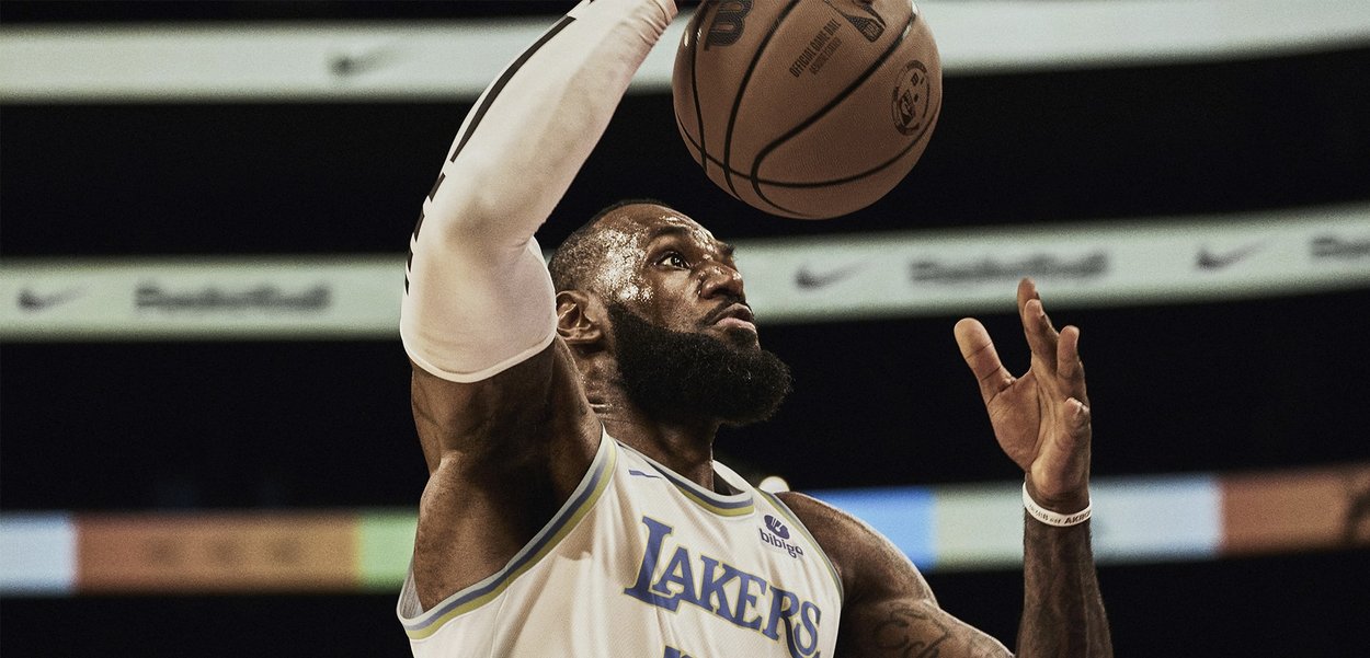 Joueur de basketball portant un maillot blanc des Lakers avec des accents bleus, bras tendu pour tirer, tenant un ballon de basket brun, avec une barbe texturée.