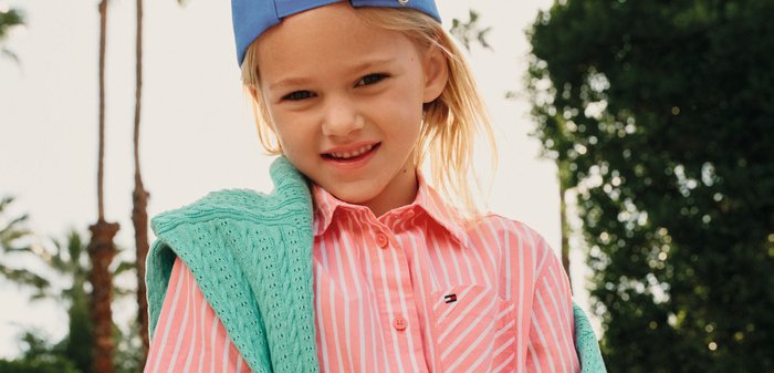 Junges Mädchen mit blauer Mütze, die rückwärts getragen wird, einem rosa gestreiften Kleid und einem grünen Pullover über den Schultern, das im Freien in der Nähe von Palmen und einem blauen Auto steht.