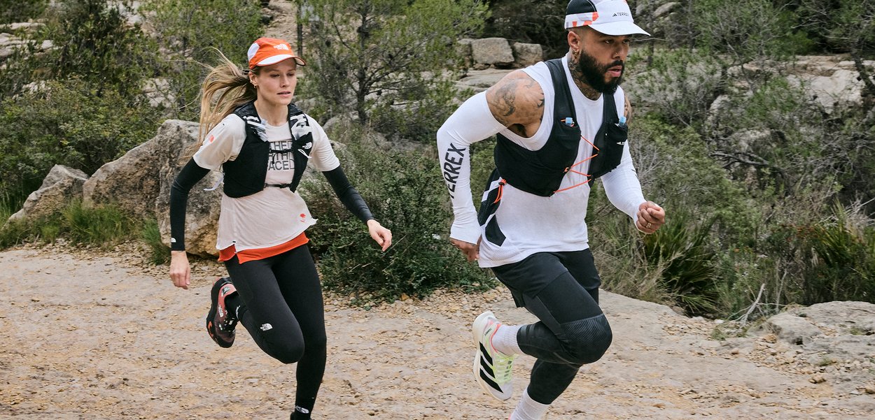 Deux coureurs de trail portant des tenues de sport et des sacs d'hydratation courent en montée sur un sentier rocheux entouré de buissons et d'arbres.