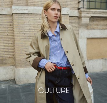 Blonde Frau mit Perlenohrringen trägt einen beigen Trenchcoat über einem blau karierte Hemd und dunklen Jeans mit rotem Gürtel, steht an einer Backsteinmauer.