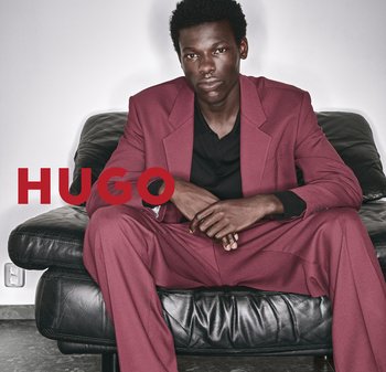 Bordeaux Anzug mit strukturiertem Blazer und weit geschnittenen Hosen, über einem schwarzen Poloshirt getragen, sitzt auf einem schwarzen Ledersofa. Rotes Logo mit der Aufschrift "HUGO."