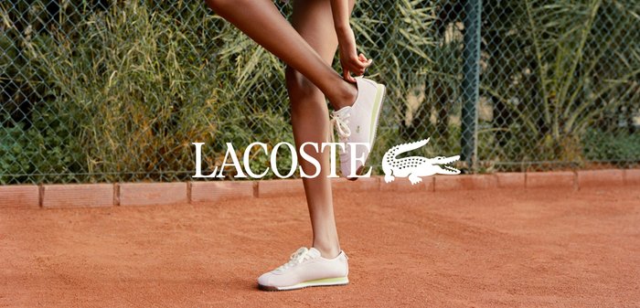 Inimene kohandab valgeid tenniseid, seistes savitenniseväljakul, kus taga on aedvõrk ja rohelus.