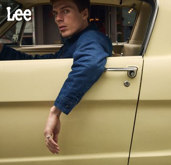 Hombre con chaqueta de mezclilla azul oscuro sentado en un coche vintage beige, con el brazo colgando de la ventana abierta, mirando por encima del hombro.