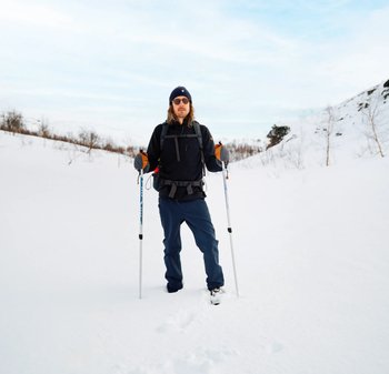 Ein Outdoor-Wanderer in schwarzer Jacke und blauen Hosen steht im Schnee und benutzt Trekkingstöcke mit orangefarbenen Handschuhen. Winterlandschaft im Hintergrund.
