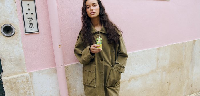 Young woman with long curly hair wearing an oversized olive coat and grey trousers, holding a juice carton, standing against a pink and beige wall.