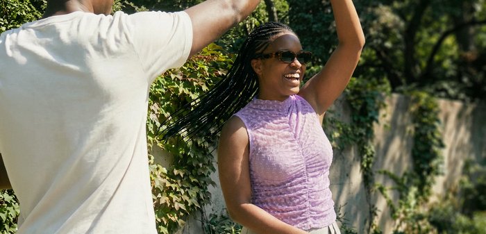 Una mujer sonriente con gafas de sol y trenzas baila al aire libre con un hombre que levanta el brazo junto a una pared verde cubierta de hiedra.