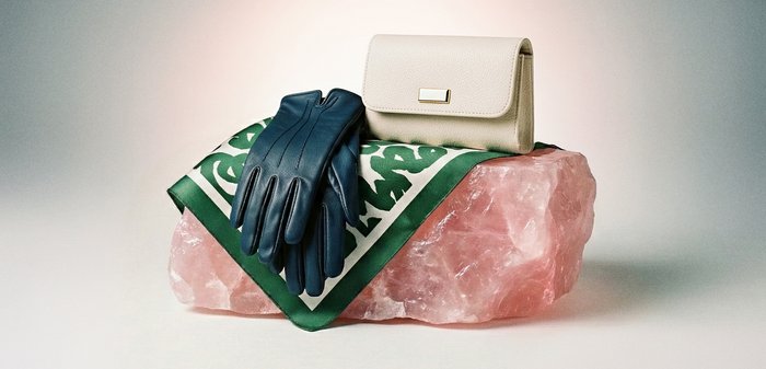 Melnas ādas cimdi un bēša krāsas maki atrodas uz zaļas un baltas šalles, kas pārklāta pāri lielam rozā kristāla akmenim.