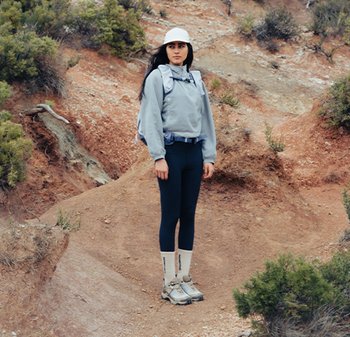 Jeune femme portant un équipement d'extérieur et une casquette, debout sur un terrain rocheux avec une végétation clairsemée, regardant sur le côté.
