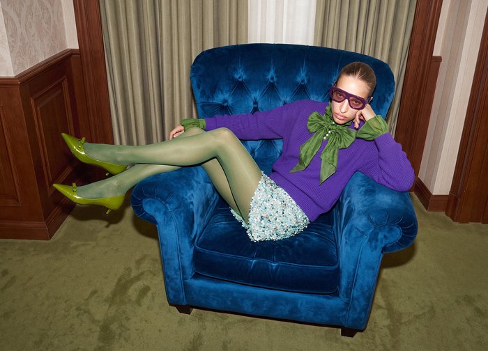 Une femme vêtue de violet, chaussée de talons verts et portant de grandes lunettes, est allongée dans un fauteuil en velours bleu, dans une pièce lambrissée.