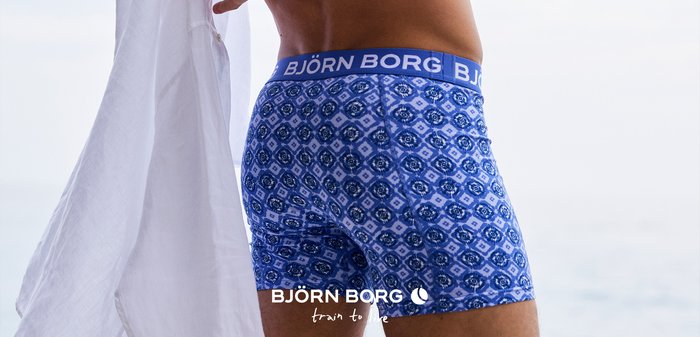 Man som bär blåmönstrade Björn Borg-boxershorts och håller en vit skjorta mot en ljus bakgrund.