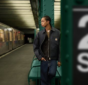 Νεαρός με ζώνες στα μαλλιά, φορώντας ένα σκούρο μπουφάν και τζιν, στέκεται σε μια αποβάθρα του μετρό κοντά σε ένα πράσινο παγκάκι, κοιτάζοντας δεξιά.