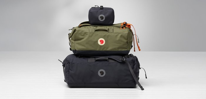 Drei Reisetaschen: eine olivgrün und zwei schwarz in unterschiedlichen Größen. Alle haben eine Stofftextur, hervorgehobene Logos und verstellbare Riemen.