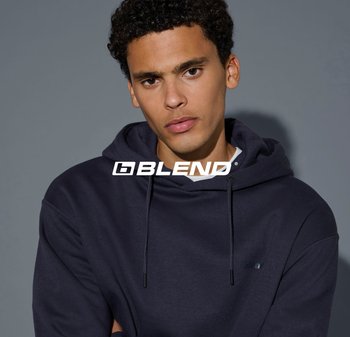 Junger Mann mit lockigem Haar, der einen dunkelblauen Hoodie mit Kordelzug vor einem schlichten grauen Hintergrund trägt.