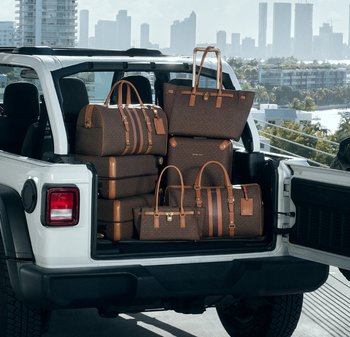 Maletero abierto de un SUV blanco cargado con equipaje y bolsos de diseño marrones, con el skyline de la ciudad visible al fondo.