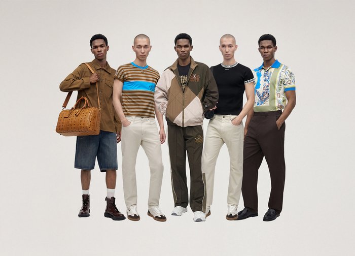 Cinq hommes se tiennent debout dans des tenues décontractées et sportives comprenant des shorts, des vestes, des chemises rayées et à motifs, des pantalons blancs et foncés, ainsi que des baskets ou des bottes.