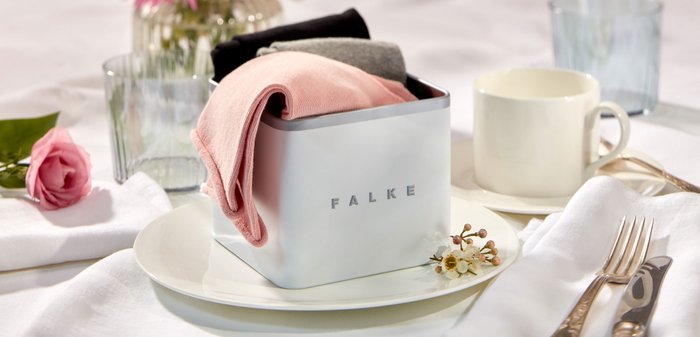 Quadratischer Metallbehälter aus gebürstetem Silber mit eingeprägtem "FALKE", der gefaltete rosa, schwarze und graue Kleidungsstücke hält, auf einem weißen Teller platziert.