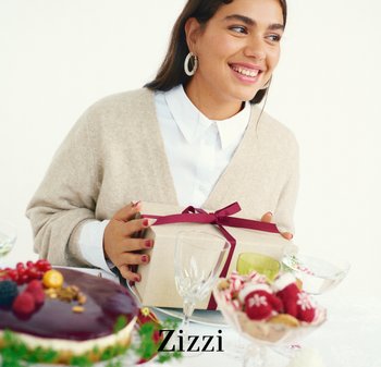 Presentkartong inslagen i beige papper med ett burgundyfärgat band, hålls av en person i en gräddfärgad kofta, med desserter och glas på bordet.