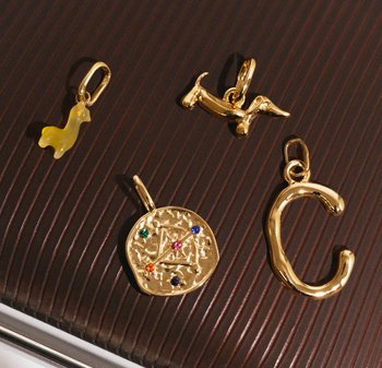 Quatre breloques dorées : un chien en résine jaune, une figurine de chien en or, un pendentif rond gravé avec des pierres précieuses multicolores, et une lettre "C" en écriture cursive.