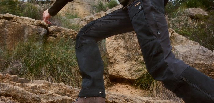 Persoon die zwarte broek en bruine laarzen draagt en een rotsachtig terrein beklimt met gras en grote rotsen op de achtergrond.