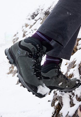 Μαύρες ορειβατικές μπότες με υφασμάτινη υφή και καουτσούκ σόλες, οι οποίες διαθέτουν κορδόνια και ενισχυμένα καπάκια δαχτύλων, μισοσκεπασμένες με χιόνι, φορεμένες με μωβ κάλτσες.