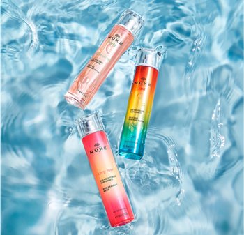 Tre bottiglie di profumo Nuxe con design cilindrico e trasparente: "Very Rose" rosa, "Sun" con gradiente vibrante e blu, tutte impostate su uno sfondo acquatico.