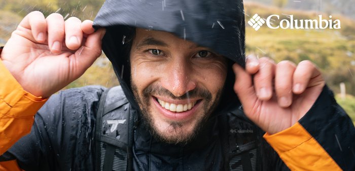 Schwarze, wasserfeste, kapuzenjacke mit einem lächelnden Mann, der die Kapuze anpasst. Die Jacke hat graue Riemen und wird über einem gelben Langarmshirt getragen.