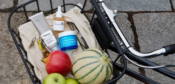 En svart metallcykelkorg håller två äpplen, en cantaloup, och tre hudvårdsprodukter i vita och blå behållare på en krämfärgad duk.