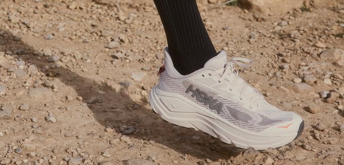 Witte Hoka hardloopschoen met zwarte sokken die op een rotsachtige, stoffige ondergrond stappen tijdens een buitenactiviteit.