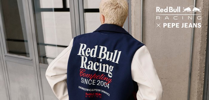 Pessoa com cabelo curto e loiro veste uma jaqueta azul-marinho e branca da Red Bull Racing com o texto "Competing since 2004", estando em interiores perto de uma janela.