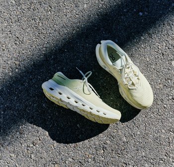 Hellbeige Sportschuhe zum Laufen mit dicken, belüfteten Sohlen, die auf grauem Asphalt im Sonnenlicht und Schatten liegen.