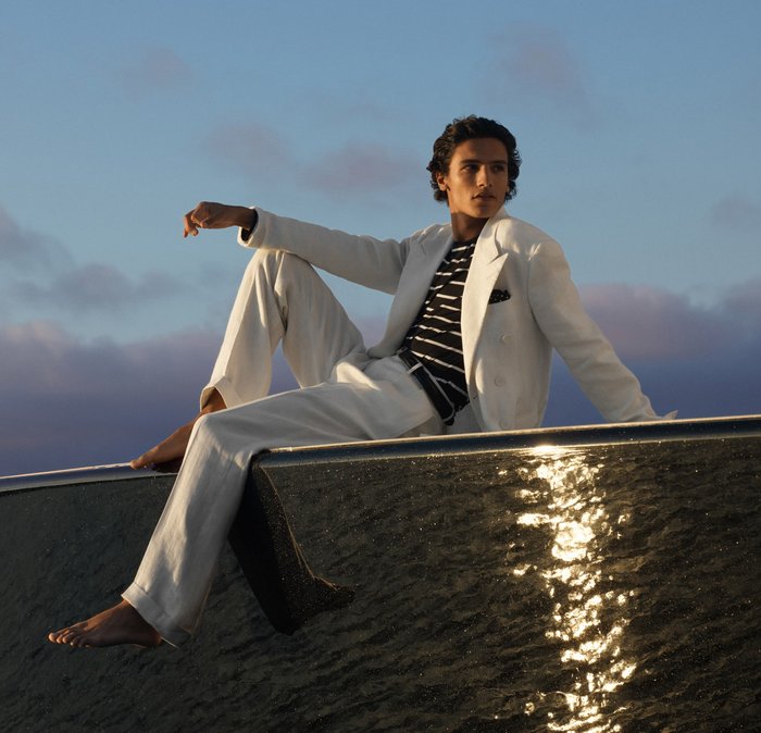 Joven vestido con traje blanco y camisa de rayas, sentado descalzo sobre una superficie negra reflectante al atardecer, con un cielo nublado de fondo.