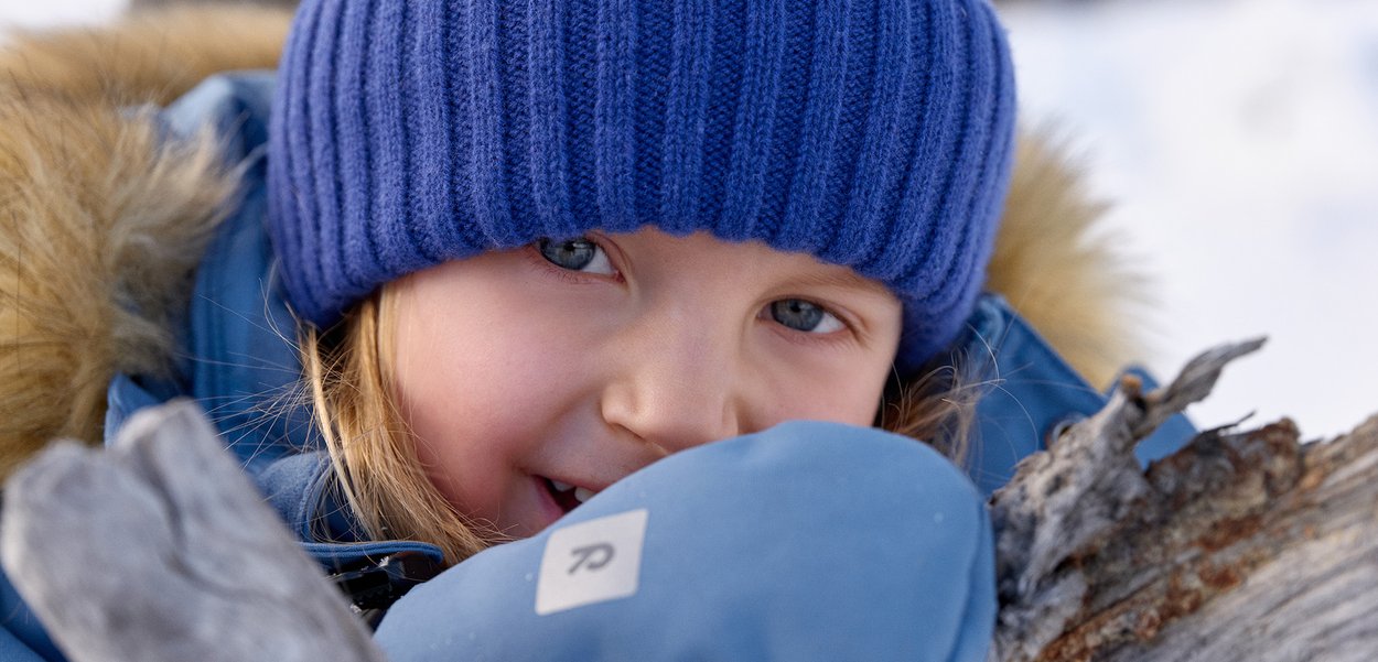 Sininen neulepipo, pehmeä pinta, yhdistettynä siniseen paksuun talvitakkiin. Lapsi hymyilee, pitäen harmaata lapasta puuoksaa vasten.