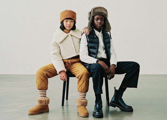 To børn sidder på en stol, den ene iført en hvid shearling-kravejakke og tan kviltede bukser; den anden i en navy puffer vest og sorte bukser.