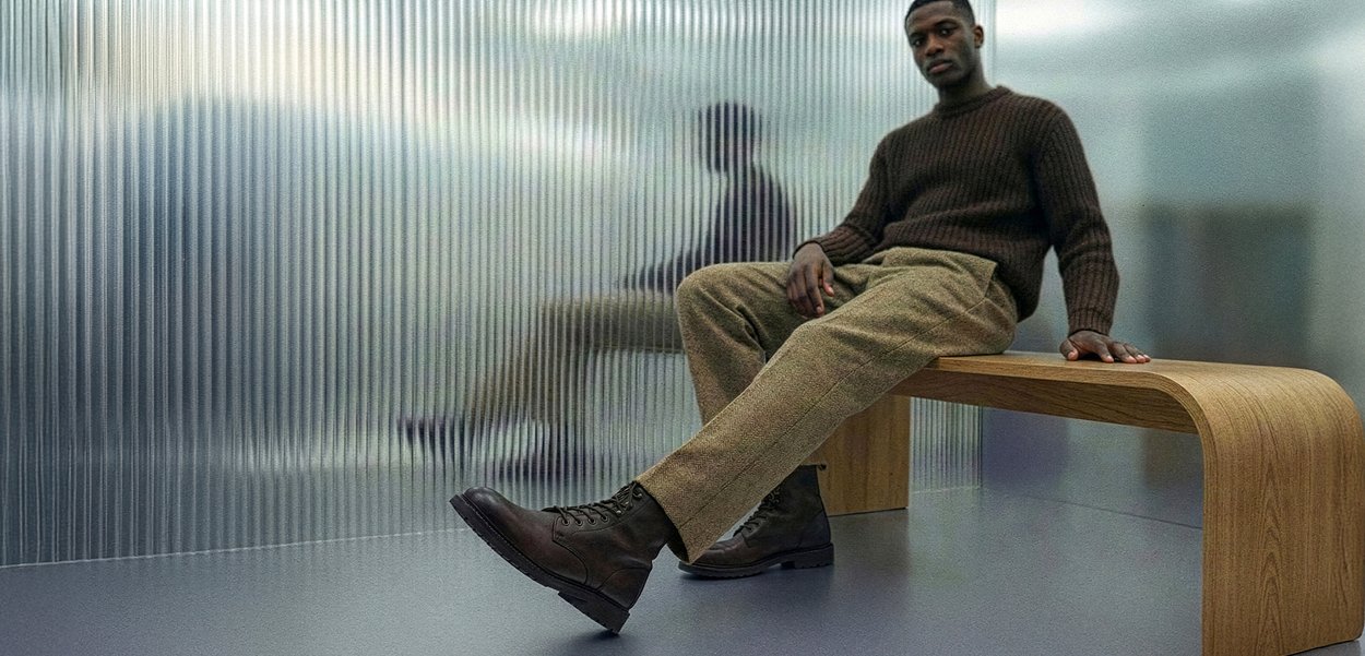 Άνδρας καθισμένος σε ξύλινο πάγκο φοράει καφέ πουλόβερ, μπεζ παντελόνι και σκούρα καφέ μπότες, με την αντανάκλασή του να φαίνεται σε υφή υαλοπίνακα πίσω του.