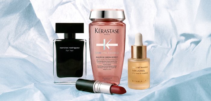 Quatre produits de beauté sur un tissu bleu clair : parfum Narciso Rodriguez, shampooing rose Kérastase, sérum Rituals et rouge à lèvres rouge foncé.