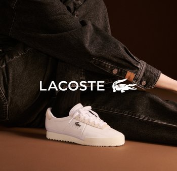 Zapatillas Lacoste blancas de cuero texturizado, con puntera redonda y cordones. Llevadas con jeans de mezclilla negros que presentan un puño abotonado y etiqueta de cuero.