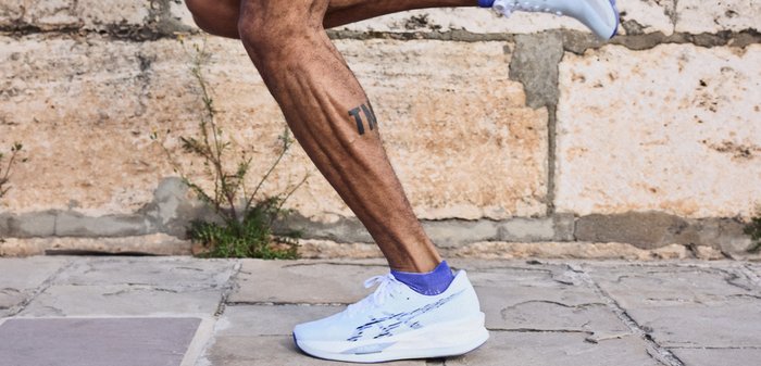 Μυώδες πόδι δρομέα με τατουάζ, φορώντας λευκά αθλητικά παπούτσια και μωβ κάλτσες, σε μέσο βήμα σε πέτρινο πεζοδρόμιο κοντά σε ένα τοίχο.